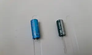 Dwa kondensatory elektrolityczne, jeden niebieski, drugi szary, z oznaczeniami wskazującymi na polaryzację kondensatora.