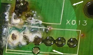 Uszkodzona płytka drukowana z widocznym śladem przepalenia i resztkami białej substancji. Widać fragmenty białych ramek i oznaczenie "X013". Zastosowano conformal coating.