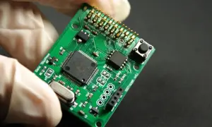 Zielona płytka drukowana z elementami elektronicznymi, trzymana w białej rękawiczce. To jest laminat, podstawa elektroniki.