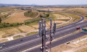 Wieża telekomunikacyjna z panelami antenowymi stoi obok autostrady. W tle rozciąga się krajobraz z polami i wzgórzami, a nad nimi widać pasmo górskie.