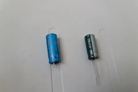 Dwa kondensatory elektrolityczne, jeden niebieski, drugi szary, z oznaczeniami wskazującymi na polaryzację kondensatora.