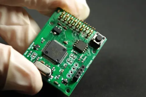 Zielona płytka drukowana z elementami elektronicznymi, trzymana w białej rękawiczce. To jest laminat, podstawa elektroniki.