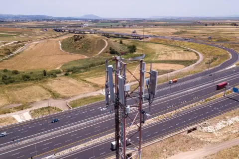 Wieża telekomunikacyjna z panelami antenowymi stoi obok autostrady. W tle rozciąga się krajobraz z polami i wzgórzami, a nad nimi widać pasmo górskie.