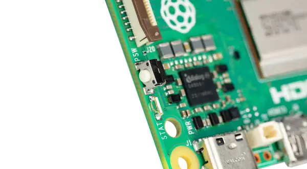 Zbliżenie na zieloną płytkę drukowaną Raspberry Pi 6 z widocznym przyciskiem, złączami i logo.