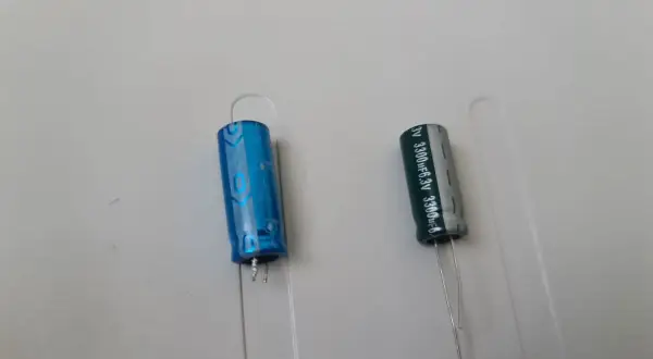 Dwa kondensatory elektrolityczne, jeden niebieski, drugi szary, z oznaczeniami wskazującymi na polaryzację kondensatora.