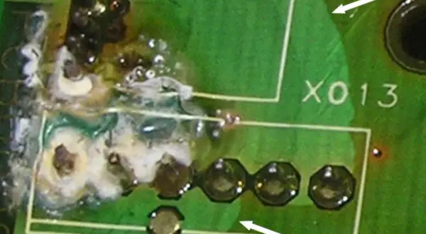 Uszkodzona płytka drukowana z widocznym śladem przepalenia i resztkami białej substancji. Widać fragmenty białych ramek i oznaczenie "X013". Zastosowano conformal coating.