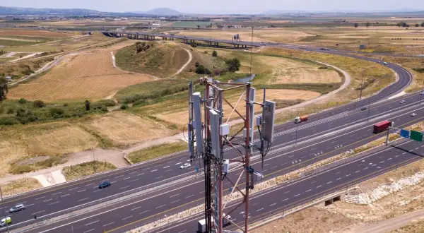 Wieża telekomunikacyjna z panelami antenowymi stoi obok autostrady. W tle rozciąga się krajobraz z polami i wzgórzami, a nad nimi widać pasmo górskie.
