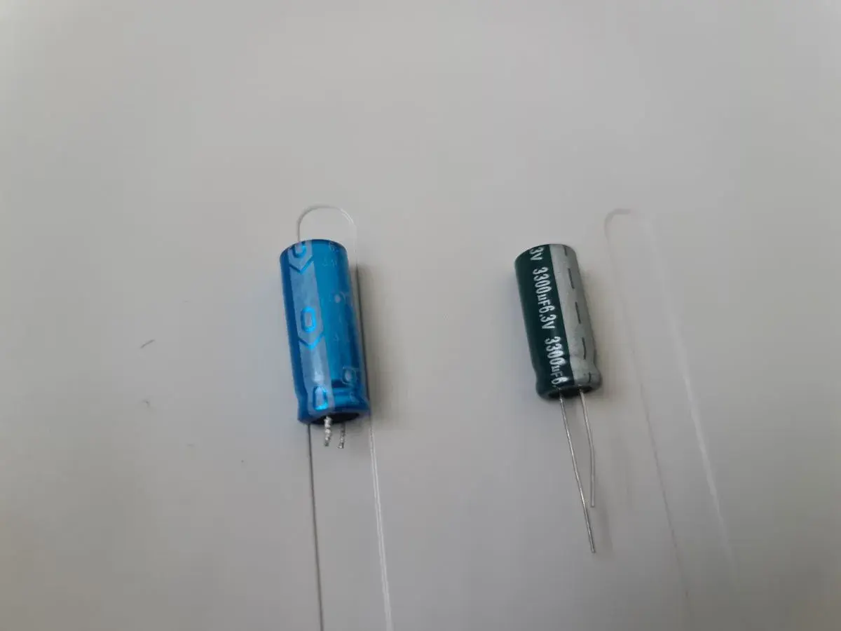 Dwa kondensatory elektrolityczne, jeden niebieski, drugi szary, z oznaczeniami wskazującymi na polaryzację kondensatora.