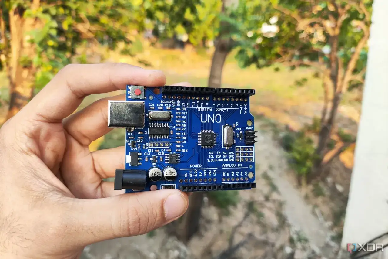 Dłoń trzyma niebieską płytkę Arduino Uno, gotową do arduino programowanie. W tle zamazane drzewa.