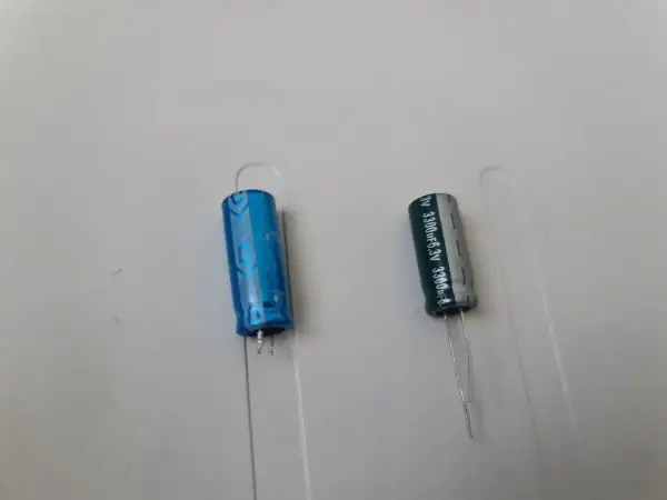 Dwa kondensatory elektrolityczne, jeden niebieski, drugi szary, z oznaczeniami wskazującymi na polaryzację kondensatora.