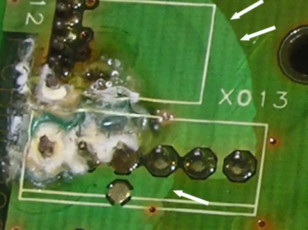 Uszkodzona płytka drukowana z widocznym śladem przepalenia i resztkami białej substancji. Widać fragmenty białych ramek i oznaczenie "X013". Zastosowano conformal coating.
