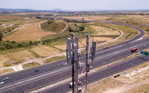 Wieża telekomunikacyjna z panelami antenowymi stoi obok autostrady. W tle rozciąga się krajobraz z polami i wzgórzami, a nad nimi widać pasmo górskie.